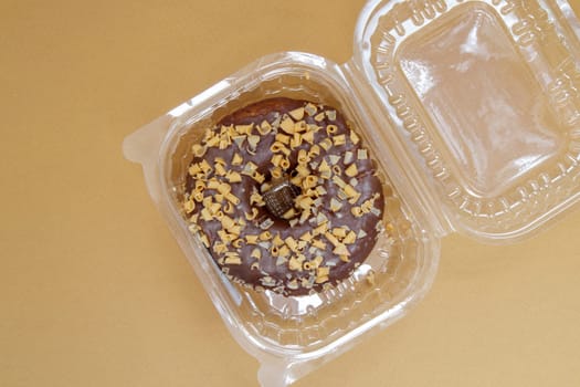 Chocolate donut in a plastic container on a brown or coffee background. Takeaway breakfast concept. One donut is packed in a plastic box for delivery. Sweet pastries delivered to your home. Top view