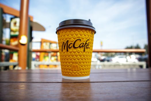 Paper yellow glass glass with coffee for McCafe drinks on the table against blurred city from McDonald's. Fast food business background. Fast food restaurant menu. Ukraine, Kiev - September 13, 2021