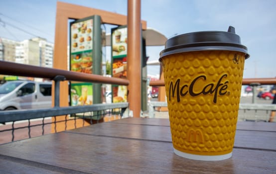 Paper yellow glass glass with coffee for McCafe drinks on the table against blurred city from McDonald's. Fast food business background. Fast food restaurant menu. Ukraine, Kiev - September 13, 2021