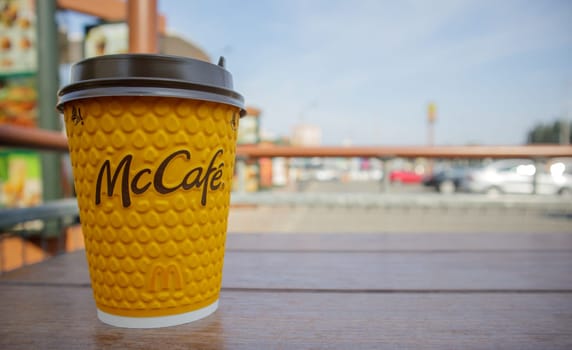 Paper yellow glass glass with coffee for McCafe drinks on the table against blurred city from McDonald's. Fast food business background. Fast food restaurant menu. Ukraine, Kiev - September 13, 2021
