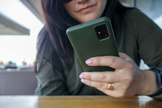Close up of woman's hand with wedding ring holding cell or mobile phone during coffee break. A woman in a green dress is reading something on her smartphone while sitting in a modern cafe