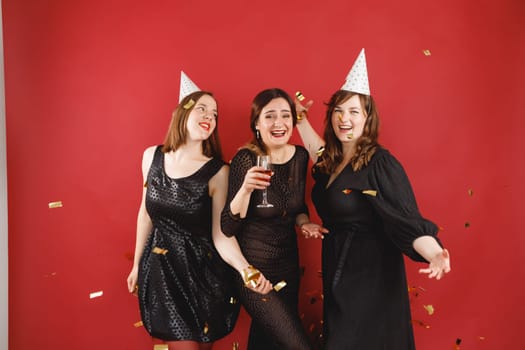 Three delighted women in black dress and party hats over red background with golden confetti. Female party