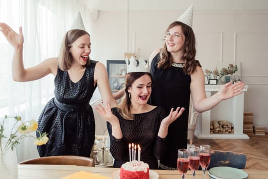 Three women friends celebrating 30s birthday together gathering at table at home with wine and birthday cake with candles. Women in party hat and birthday girl in crown blows out candles
