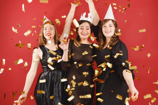 Three delighted women in black dress and party hats over red background with golden confetti. Female party