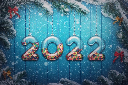 Happy New Year 2022 holiday background. Set of transparent numbers made of glass filled with multi colored candy and sweets hang on snowy Christmas tree branches as decoration garlands.