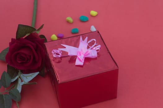 Close up gift box with tied bow ribbon placed near red rose on a table near in red background. High angle view. Design for valentine's day, anniversary or holiday celebration decoration.