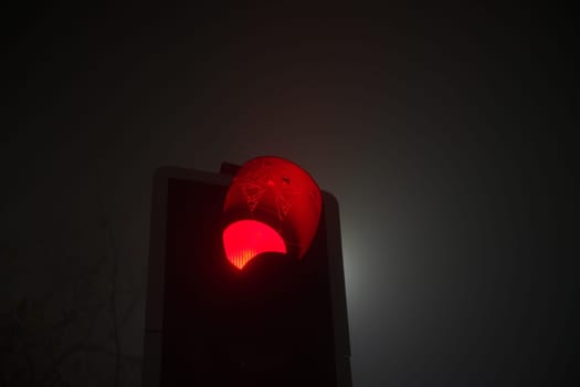 A british traffic light shines red, indicating stop, during foggy conditions at night. Also, a cobweb hangs over the red light of the traffic signal