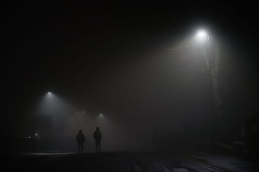 Two mysterious hooded men walk away down a dark foggy road, lit by dim white street lamps. Both figures wear coats and walk with hands in pockets.