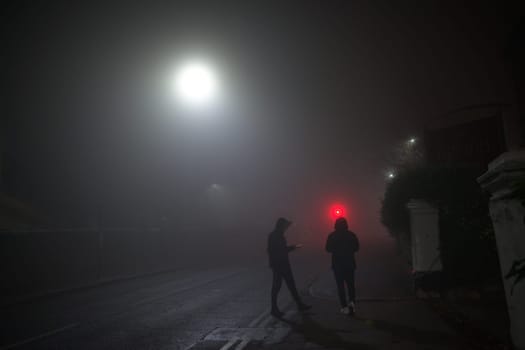 Hooded youths loiter on a street corner using mobile smartphones. Dark and foggy conditions with urban setting. Both wear winter clothes with trainers