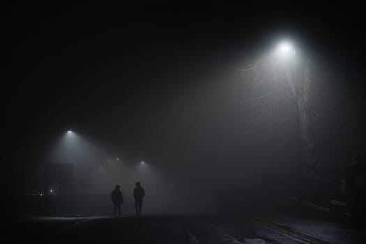 Urban winters scene of two men walking under street lights in extremely foggy conditions. Moody or spooky feel, both wear black coats with hoods up.