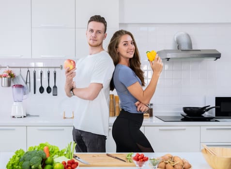Smiling couple standing in kitchen while preparing healthy food. Man holding red apple and woman holding yellow bell pepper.