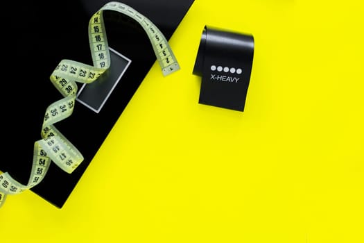A weight scale, a black fitness tape and a yellow measuring tape lie on a yellow background on the side of the photo.