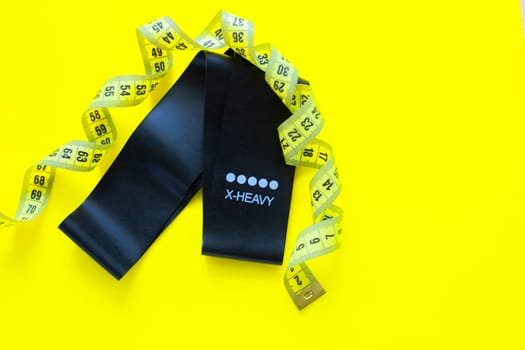 A black fitness elastic band lies on a yellow background, it is wrapped around a yellow measuring tape. top view, flat lay, copy space, isolate. High quality photo