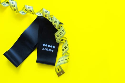 A black fitness elastic band lies on a yellow background, it is wrapped around a yellow measuring tape. top view, flat lay, copy space, isolate. High quality photo