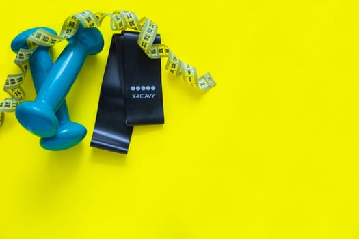A black fitness elastic band and two blue dumbbells lie on a yellow background, a measuring tape lies above them in rings. top view, flat lay, copy space, isolate. High quality photo
