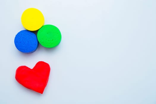 A heart made of red dough for modeling lies on top of the photo, and yellow, green and blue circles lie at the bottom on a white background.. High quality photo