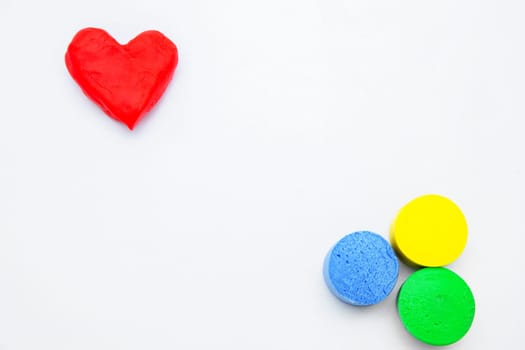 A heart made of red dough for modeling lies on top of the photo, and yellow, green and blue circles lie diagonally on a white background. High quality photo
