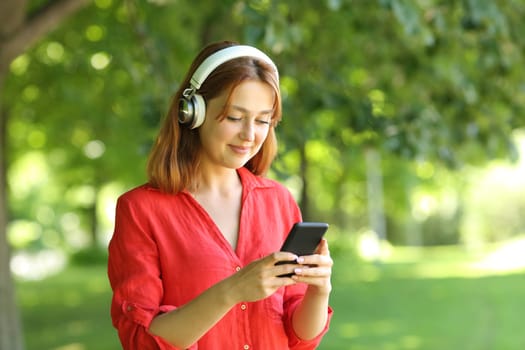 Happy woman in red wearing wireless headphones is waling listening to music on smartphone in a park