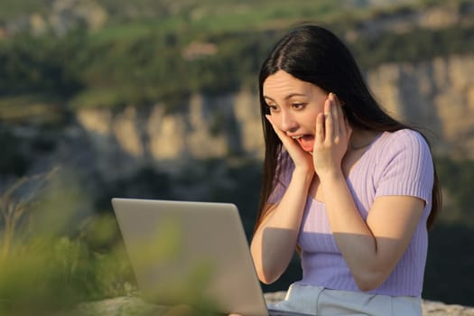 Excited asian woman watching laptop media content sitting in nature