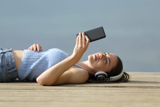 Serious woman lying on pier listening to music with smart phone