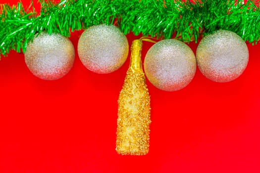Christmas balls of silver and a golden bottle of champagne hang on a green fluffy garland of tinsel green on top of the photo on a red background. top view, flat lay, copy space, isolate. High quality photo