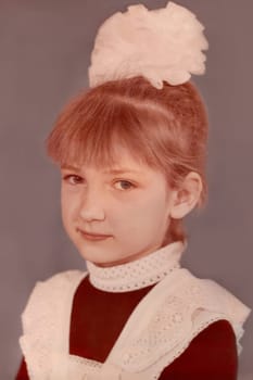 First-grader girl, seven years old, with a bow on her head and in a school uniform, white apron. Vintage photo from 1988. USSR