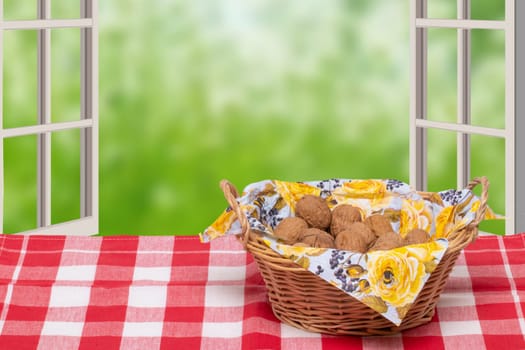 A wicker basket with a napkin full of walnuts on a table with a red checkered tablecloth in front of open windows with an abstract blurred natural green background. Macro.