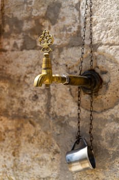 closeup of vintage bronze water faucet with decorated tap and metal drinking cup next to a Muslim mosque with stone background