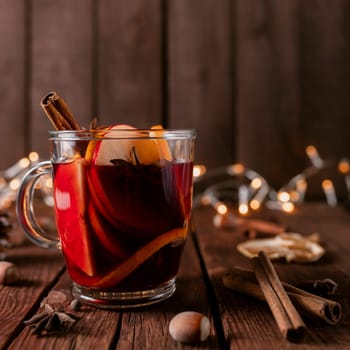 Christmas time concept photo. Glass cup or mug with Mulled Wine inside on wooden table background. Garlands, decorations, cinnamon stick, hazelnut, dried or sublimated orange, apple, various spices
