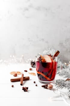 Christmas time concept photo. Glass cup or mug with Mulled Wine on white gray table background. Garlands and lights, Christmas decorations, cinnamon, orange, apple, spices, white fir, cone, anise