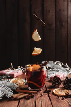 Christmas glass cup or mug with Mulled Wine on wooden table and background with levitating cinnamon stick, dried or sublimated orange, apple slices, present gift boxes, garlands, decoration lights