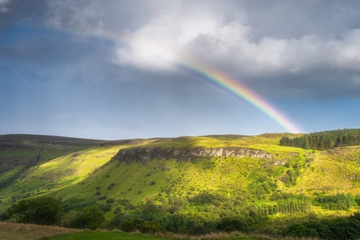 Vivid, beautiful rainbow arching over hills and forests covered in patches of sunlight, Glenariff Forest Park, County Antrim, Northern Ireland