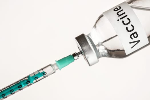 Glass vial with label Vaccine on white background, green hypodermic syringe needle inside - vaccination concept.