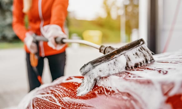 Young woman cleaning her car in self serve carwash. Closeup detail on white soap foam and brush moving over roof.