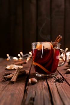 Christmas time concept photo. Glass cup or mug with Mulled Wine inside on wooden table and background. Garlands, decorations, cinnamon, hazelnut, dried or sublimated orange, apple, various spices