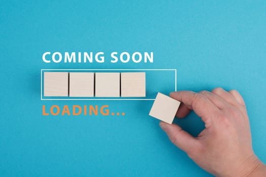 The words coming soon are standing over a loading bar, hand puts the last wooden cube to the line, blue colored backgroun
