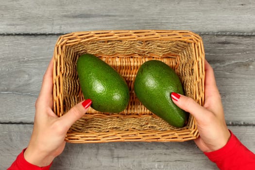 Table top view - young woman hands, red clothing and nails, holding square basket with two avocado, on gray wood desk.