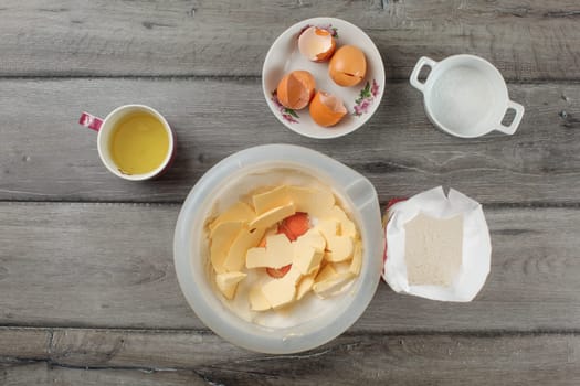 Tabletop view - plastic bowl with flour, egg yolks and butter, more ingredients around on gray wood desk. Cake preparation.