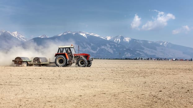 Tractor pulling heavy metal roller over dry field on a nice spring day with mountains in background. Soil preparation.