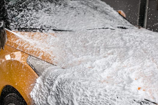Dark yellow car being washed in carwash. Front completely covered in thick shampoo foam, with more spraying from the brush.