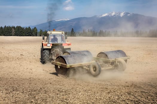 Tractor pulling heavy metal roller on dry field, dust behind and black smoke above, mountains in back. Spring ground preparation.