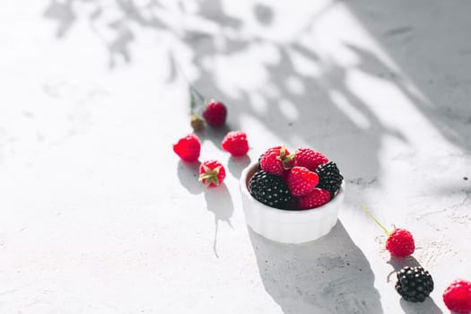 White bowl full with raspberries, blackberry on gray concrete table with leaves, tree branches shadow. Healthy eating concept. Eco, bio farming. Fresh tasty berries on grey background. Quality photo