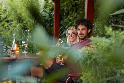 Drinking wine, couple sitting on an outdoor terrace, shot through brances of a green bush in the forground