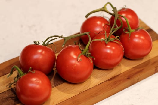 On a kitchen counter are 7 red vine ripened tomatoes with green stems on a dark and light wooden cutting board. Red tomatoes with green vine tomatoes on a dark and light wood cutting board close-up..