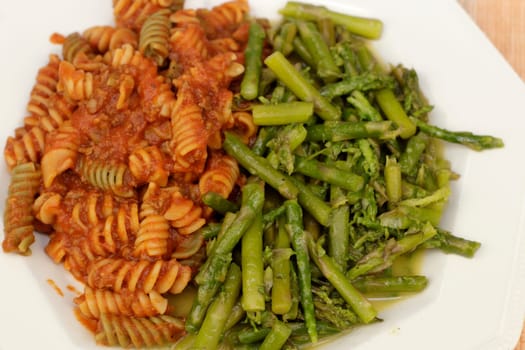 Close-up of ready-to-eat food meal of vegetable rotini pasta with tomato meat sauce with green asparagus. Cooked asparagus with butter alongside chickpea vegetable rotini pasta and meat sauce.
