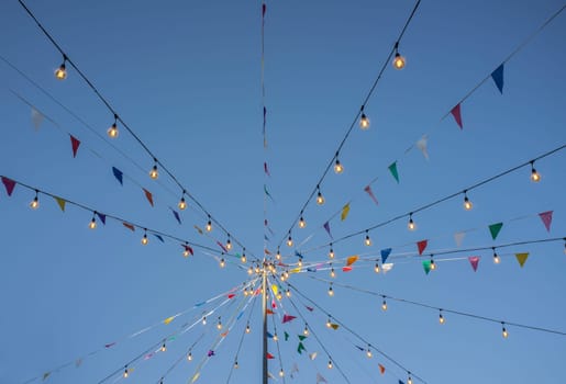 Garland with colorful pennants and light bulbs on pole over blue sky. Outdoors party background