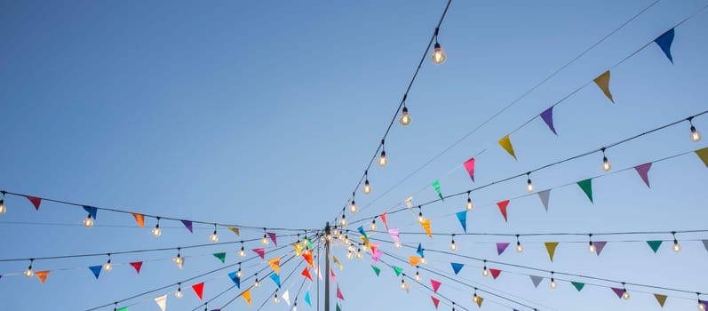 Garland with colorful pennants and light bulbs on pole over blue sky. Long format