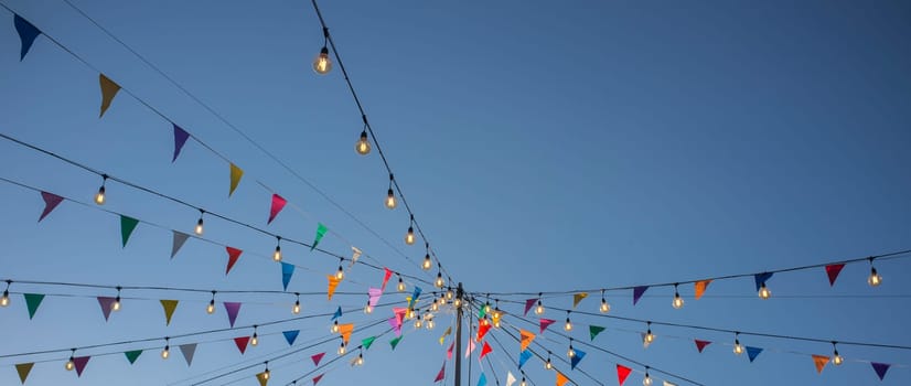 Garland with colorful pennants and light bulbs on pole over blue sky. Long format