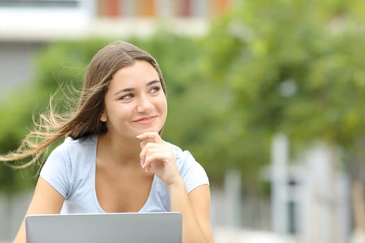 Happy teen with a laptop thinking looking at side in a park with copy space