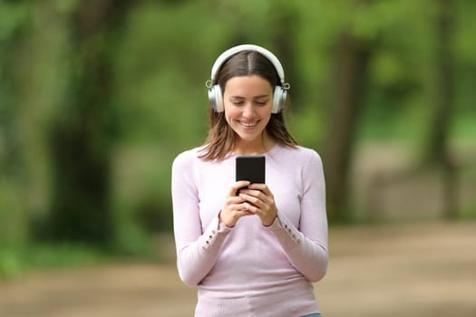 Front view portrait of a happy woman wearing wireless headphones walking listening to music on phone in a park